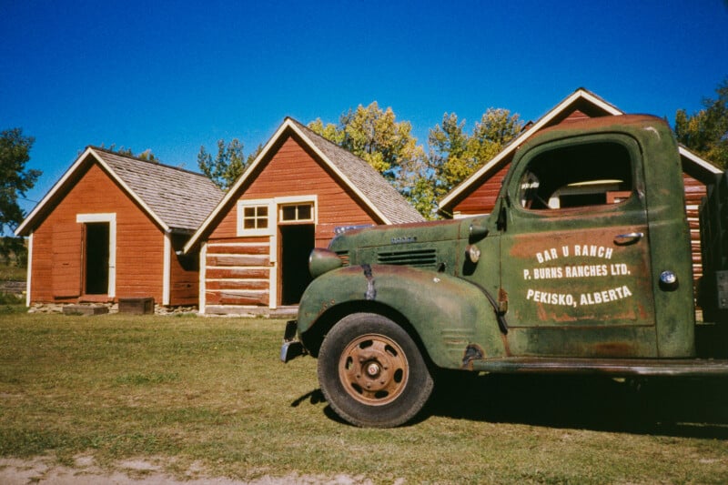 A vintage green truck is parked beside three rustic, red wooden cabins under a clear blue sky. The truck has faded lettering for "Bar U Ranch, Pekisko, Alberta" on its door.