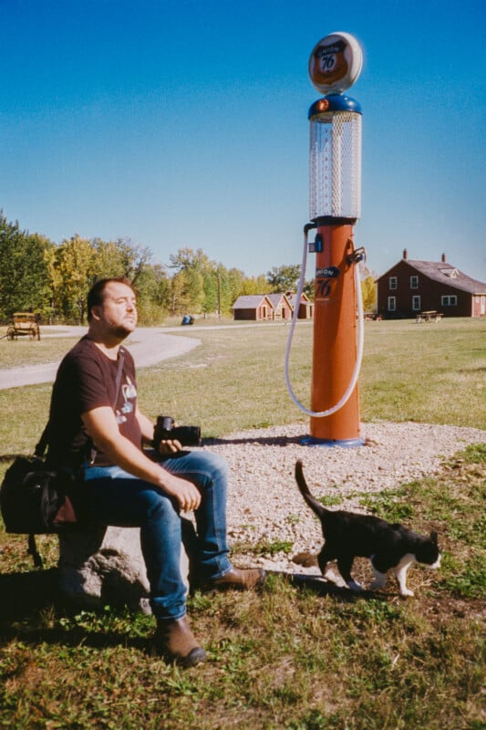 A man sits on a bench in a grassy field, holding a camera and enjoying the sun. A black and white cat walks in front of him. An old-style gas pump stands nearby, and buildings are visible in the background under a clear blue sky.