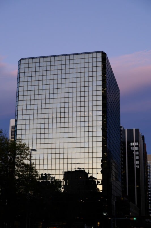 A modern glass building reflecting the twilight sky and surrounding structures. The building's grid pattern creates a geometric effect, with a hint of pinkish clouds above and cityscape elements in the reflection.