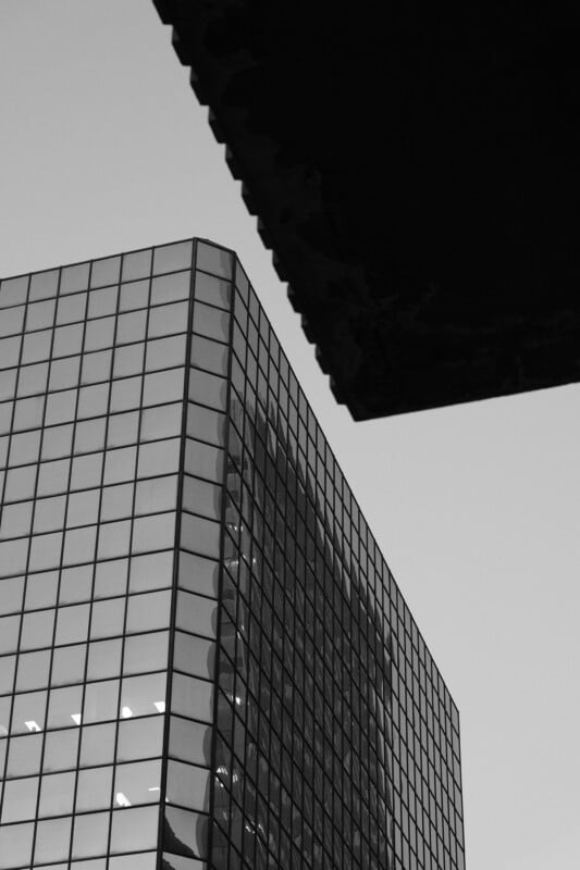 Black and white image of a modern glass office building with grid-like windows reflecting light. The corner of another structure is visible in the top right, contrasting against the clear sky.