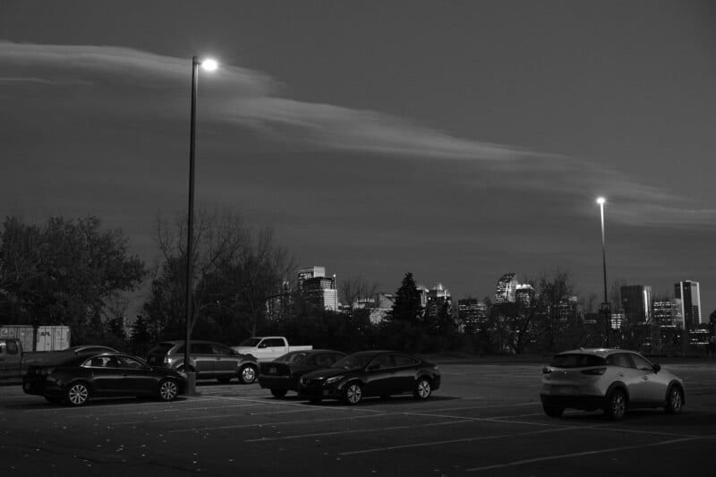 Black and white image of a parking lot with parked cars, illuminated by streetlights. In the background, a city skyline is visible under a dramatic cloud formation in the evening sky.