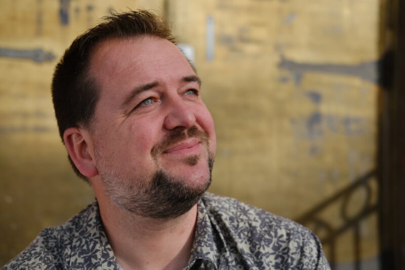 A man with short dark hair and a beard, wearing a patterned shirt, is smiling and looking upward. The background is a blurred golden surface.