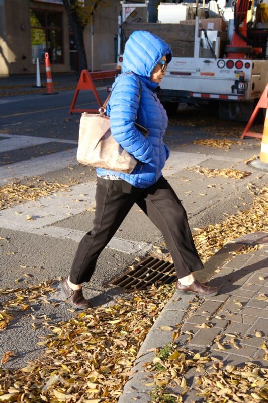 A person wearing a bright blue puffer jacket and sunglasses walks across a leaf-covered street. They carry a beige bag and are stepping onto a curb. A truck and orange construction cones are in the background.