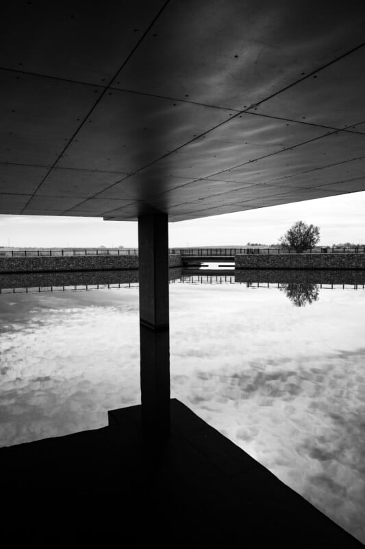 Black and white image of a modern concrete structure reflecting in a calm pool of water. A single column supports the roof, and a distant tree is visible across the water. The sky is overcast, creating a mirror-like effect on the water's surface.