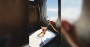 A person's hand stirring a drink with ice on a tray table by an airplane window. Sunlight casts a shadow of the glass, and fluffy clouds are visible outside against a blue sky.