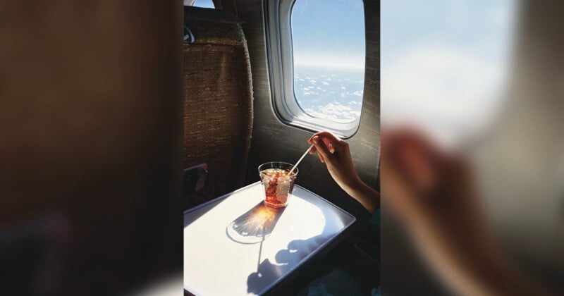 A person's hand stirring a drink with ice on a tray table by an airplane window. Sunlight casts a shadow of the glass, and fluffy clouds are visible outside against a blue sky.