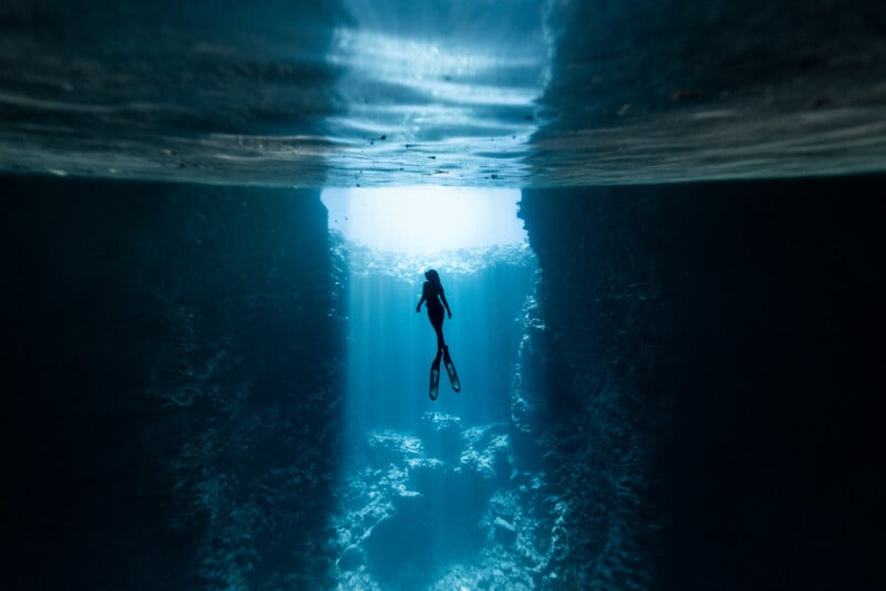 A lone diver swims through a deep, underwater canyon. Sunlight filters down from above, illuminating the clear, blue water and highlighting the rocky walls of the canyon. The serene atmosphere suggests exploration and adventure.