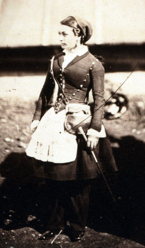 A woman in 19th-century attire wears a dark dress with a fitted jacket, apron, and cap. She holds a parasol and stands on a dirt path, looking to the side. The background is blurred.