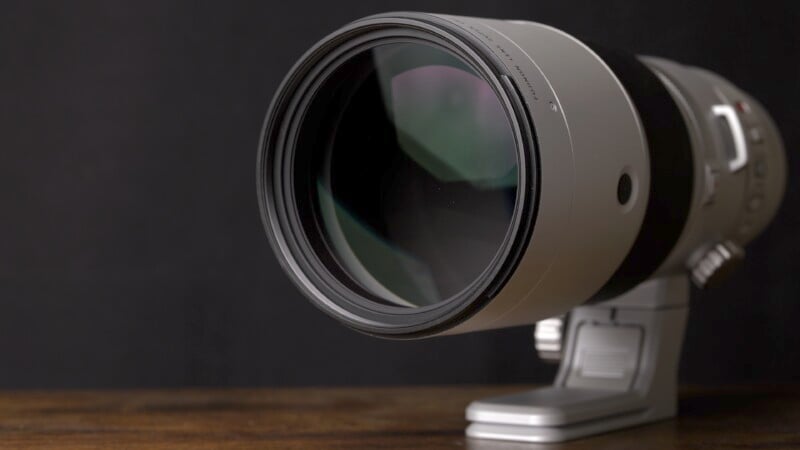 A large telephoto camera lens with a silver body and black details is mounted on a tripod collar, placed on a wooden surface against a black background.