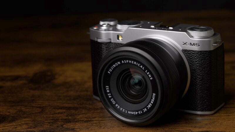 A silver and black digital camera with a Fujinon aspherical lens sits on a wooden surface against a dark background. The camera is slightly angled, showcasing its lens and textured body.