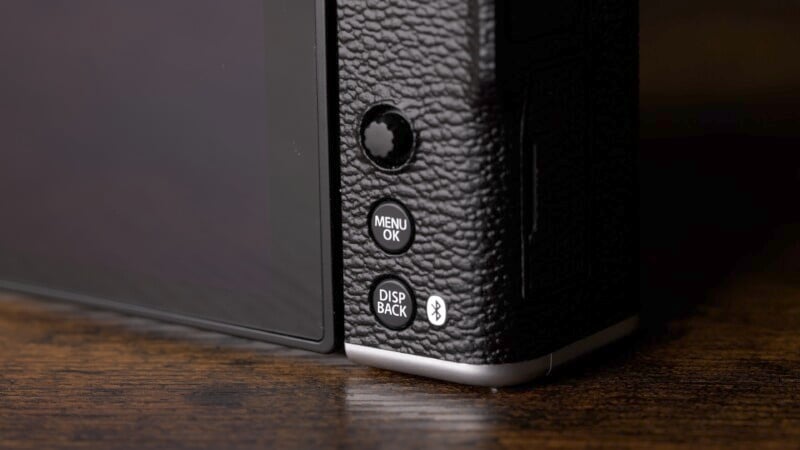 Close-up of a textured black camera with a side view of its buttons. Visible buttons include "MENU OK," "DISP BACK," and a Bluetooth symbol. The camera is placed on a wooden surface.