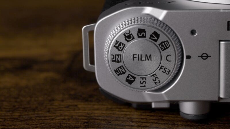 Close-up of a camera mode dial labeled "FILM," with various settings indicated. The dial is silver with a textured grip, set against a dark wooden background.