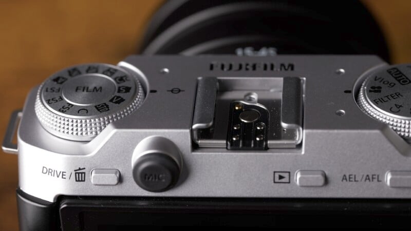 Close-up of a silver Fujifilm camera showing its control dials and buttons. The top buttons are labeled "FILM," with dials for adjusting settings. The camera's hot shoe and part of the lens are also visible.