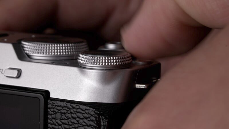 Close-up of fingers adjusting the settings on a silver camera, focusing on the dials and textured surface. The camera has a sleek design with a black grip and metallic finish.