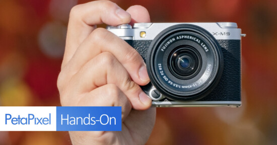 A person holding a Fujifilm X-M5 camera with an aspherical lens against a blurred background of red and orange hues. The image includes the PetaPixel logo and the text "Hands-On" at the bottom left.