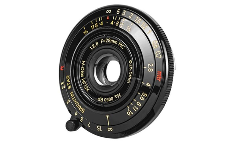 A close-up of a black circular camera lens with various markings and numbers in yellow and red, including aperture and focal length details (f/2.8, 28mm). It features a unique, compact design.