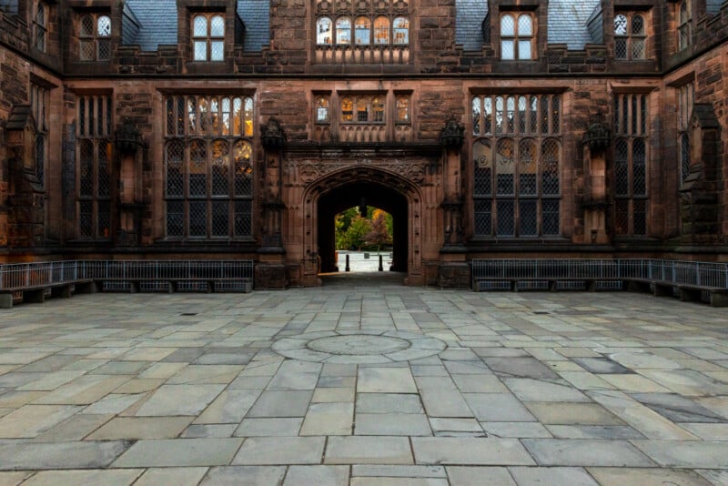 Gothic-style architectural structure with detailed stone carvings and arched doorways. The paved courtyard leads to an open archway revealing a glimpse of greenery beyond.