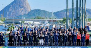 A large group of people stands on a platform, clapping, with a marina and a large mountain in the background. The stage banner reads "Global Alliance Against Hunger and Poverty." The scene is sunny and clear.