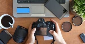 Hands hold a camera in front of a selection of other photography gear on a desk.