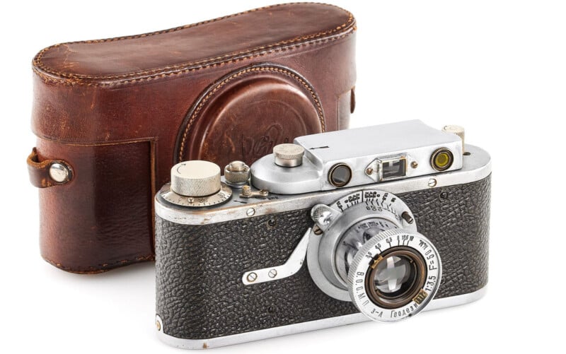 A vintage camera with a black textured body and silver metal parts is displayed in front of a brown leather carrying case. The camera features a lens with engraved details and a viewfinder on top.