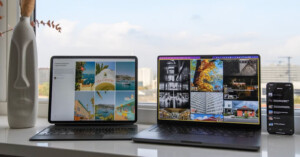 Two laptops and a smartphone are displayed on a white desk by a window. The laptops show colorful images, while the phone displays text. A vase with dried flowers is on the left. Outside, city buildings and a cloudy sky are visible.