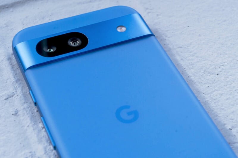 Close-up of a blue smartphone on a light surface, featuring a single rear camera and a small flash. The phone has a sleek, minimalist design with a matte finish and a subtle logo on the back.