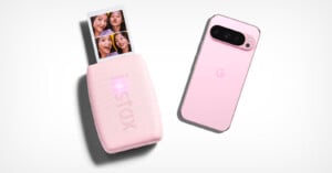 A pink smartphone with a "G" logo lies next to a compact pink photo printer. The printer is producing a strip of photo booth–style images featuring smiling faces. Both devices are set against a white background.