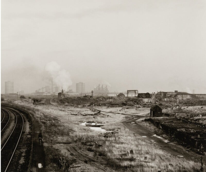 Black and white image of an industrial landscape with railway tracks on the left, open grassy fields, and distant factories emitting smoke under a cloudy sky. The scene conveys a sense of desolation and industrial activity.