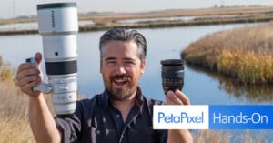 A smiling man outdoors holds a large camera lens in one hand and a smaller lens in the other. He stands near a water body with grass and reeds. The image includes a "PetaPixel Hands-On" logo.