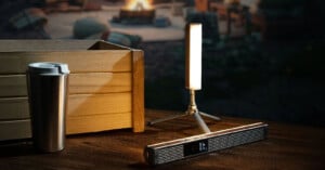 A portable LED lamp on a tripod stands on a wooden surface next to a silver travel mug and a rectangular metal object. In the background, there's a blurred image of a campfire scene with outdoor seating.