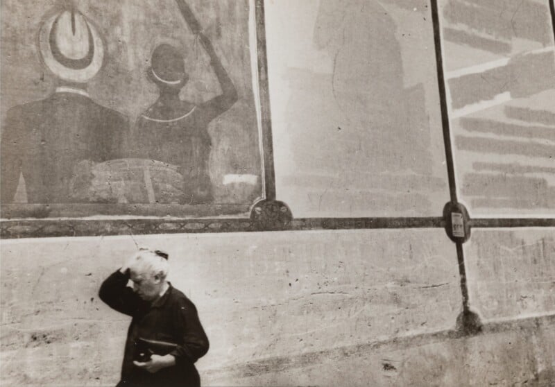 A person with tousled hair stands in front of a weathered wall decorated with faded murals depicting figures in hats. The person gazes upward, holding an object in one hand. The ground is textured, and the overall scene appears vintage.