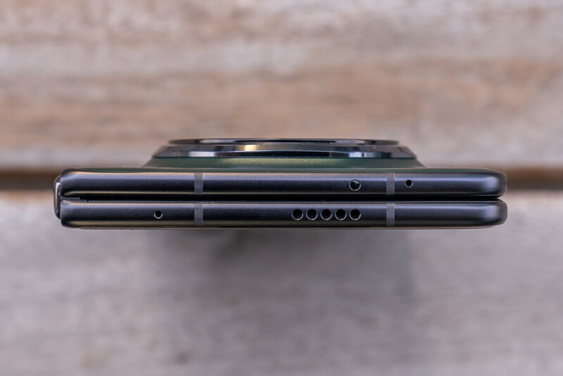 Close-up of two stacked smartphones viewed from the top edge. Both devices are black with visible side buttons and port openings. The background is a blurred wooden surface.