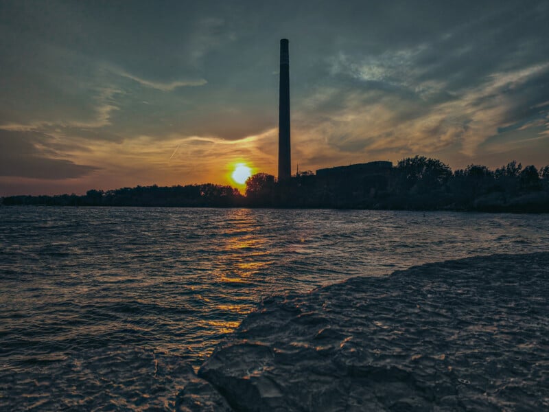 A serene sunset over a body of water, with an industrial chimney silhouetted against the sky. The sun is partially hidden by trees on the horizon, casting a warm reflection on the rippling surface of the water.