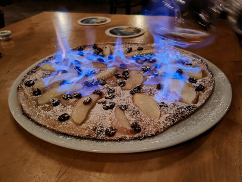 A dessert pizza topped with apple slices, raisins, and powdered sugar is flambéed, with blue flames visible over the surface. It is served on a white plate placed on a wooden table.