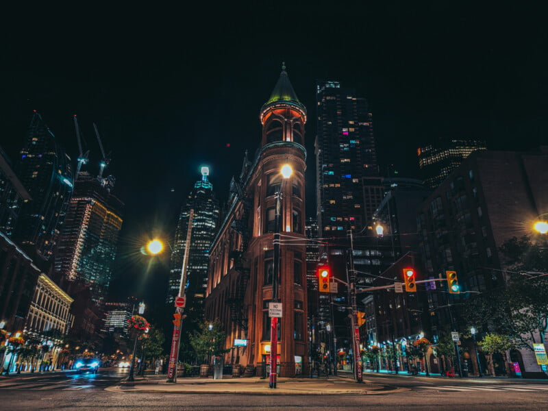 A vibrant cityscape at night, featuring an illuminated historic building with a pointed tower in the foreground. Skyscrapers light up the background, and traffic lights add color. The scene captures urban life and architecture.