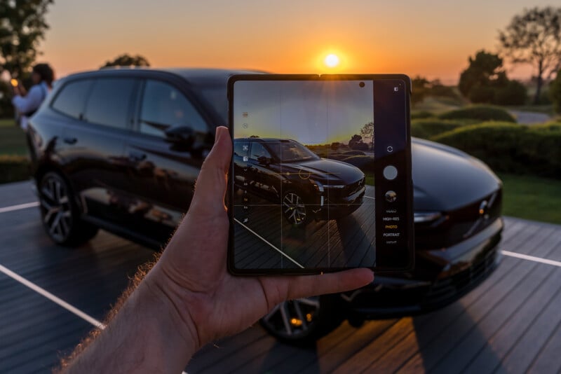 A person holds a foldable smartphone capturing a black SUV parked on a platform at sunset. The background features a scenic landscape with trees and a clear sky, reflecting the car and surroundings on the smartphone's screen.