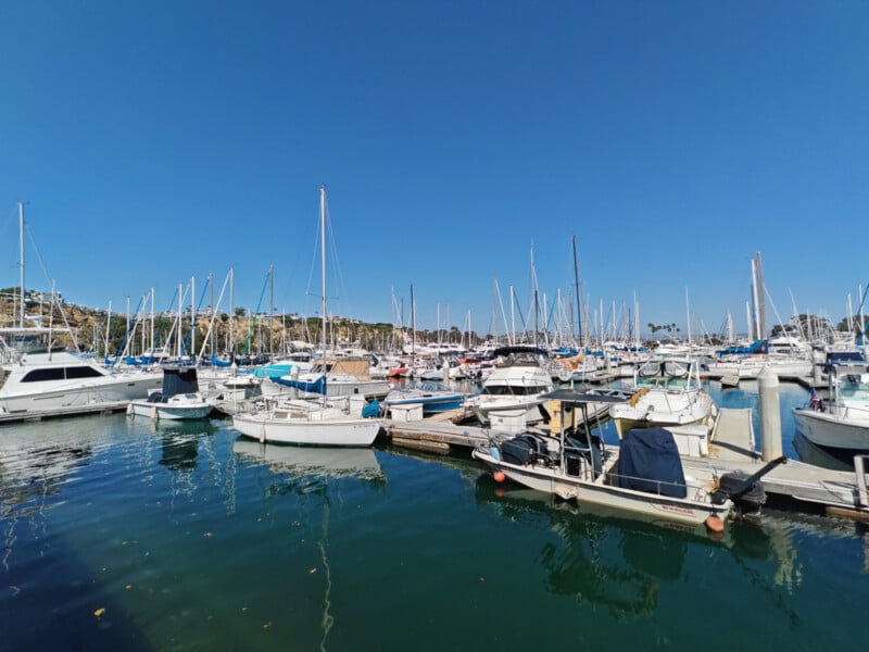 A marina filled with various sailboats and yachts docked along the piers under a clear blue sky. Hills with greenery are visible in the background. The water is calm, reflecting the boats and vibrant sky.