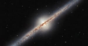 The image shows a spiral galaxy viewed edge-on against a backdrop of stars. The galaxy has a bright central bulge with a thin, elongated disk of dust and gas stretching across it, displaying some dark lanes and bright star regions.