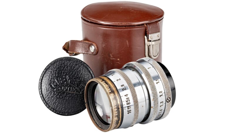 A vintage camera lens with a silver body and engraved numbers is displayed alongside a brown leather case with a buckle. A black lens cap is also shown. The scene has a clean white background.