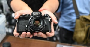 A person holds a vintage Nikon film camera towards the lens. The camera's body is black with visible details, including the Nikon logo and lens information. The background is slightly blurred, focusing attention on the camera.