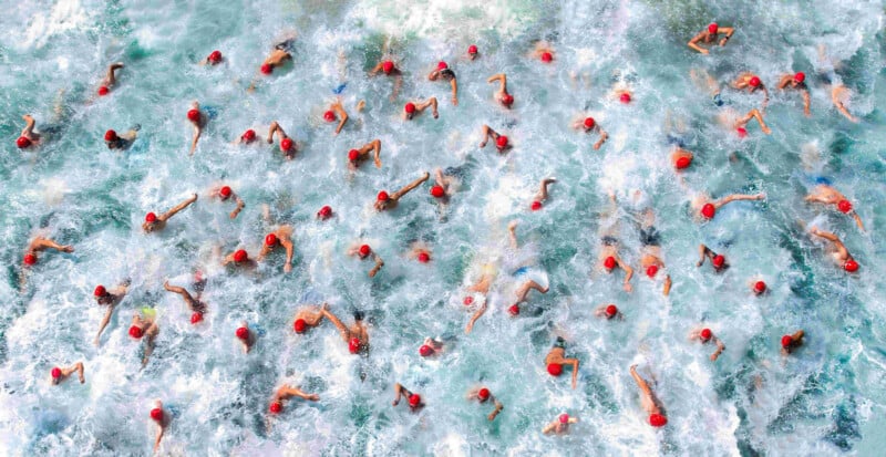 Aerial view of a large group of swimmers wearing red swim caps in turbulent water, creating a dynamic pattern of splashes and waves. The swimmers are in various swimming strokes, generating a sense of movement and energy.