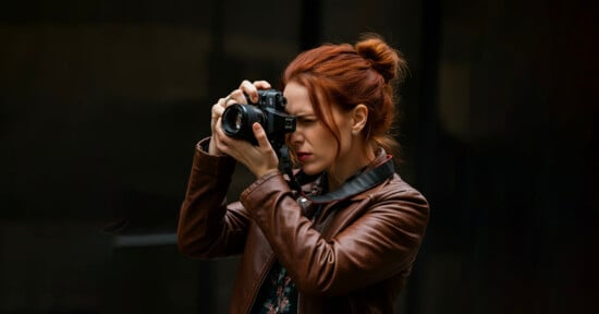 A person with red hair tied back takes a photo with a black camera. They are wearing a brown leather jacket and are focused on their photography. The background is dark and blurred.