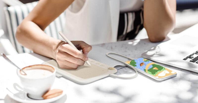 A person is using a stylus on a digital notepad at a table. Next to them is a phone with a colorful case and an open laptop. A cup of coffee sits nearby. Sunlight casts shadows on the white surface.