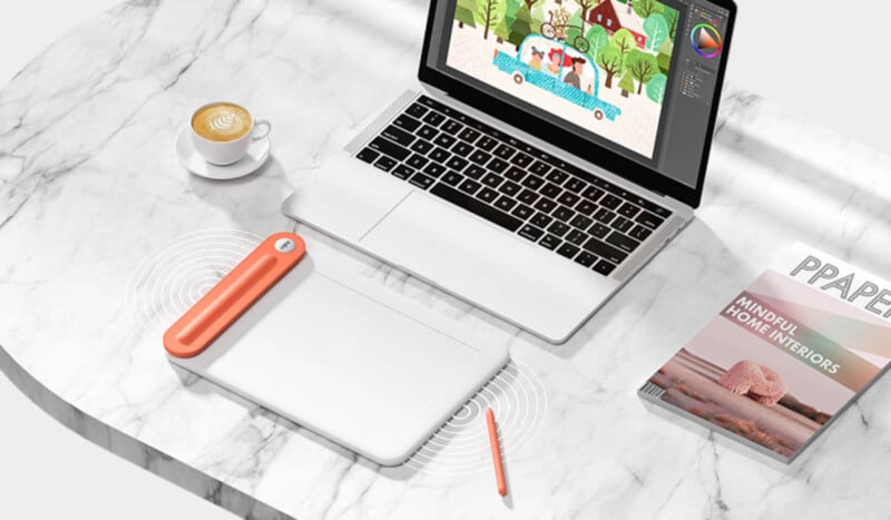 A marble table with a laptop displaying digital art, a stylus tablet, an orange stylus pen, and a cappuccino in a white cup. A magazine titled "Mindful Home Interiors" lies nearby.