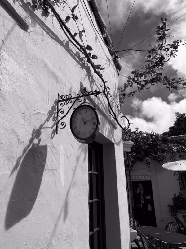 A black and white photo shows a vintage clock mounted on the exterior wall of a building. The wall is adorned with creeping vines and is partially illuminated by sunlight. The scene includes chairs and tables in an outdoor setting with a cloudy sky overhead.