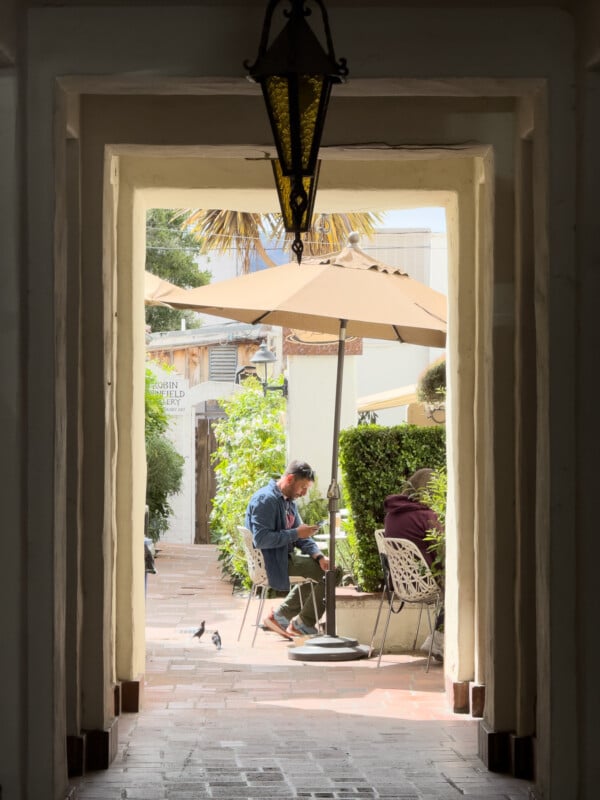 A man sits and reads at an outdoor café table under a beige umbrella, surrounded by green plants and hedges, with a hanging lantern framed by a passageway in the foreground. Sunlight illuminates the scene, creating a relaxed and peaceful atmosphere.