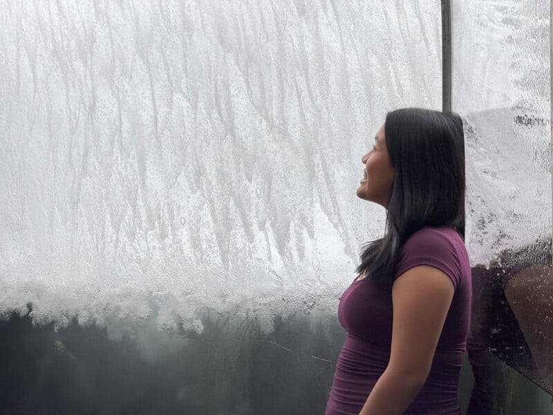 A woman with long dark hair, wearing a purple shirt, stands smiling while looking to her left. She is next to a foggy glass wall with water streaming down, creating a textured, wavy background.