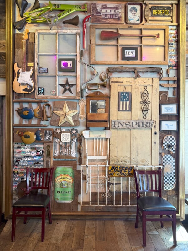 A wooden wall decorated with an eclectic mix of items including a guitar, paddle, windows, signs, a framed picture, and various knick-knacks. Two red wooden chairs sit in front, while the wall features signs that say "Inspire" and "No Trespassing.