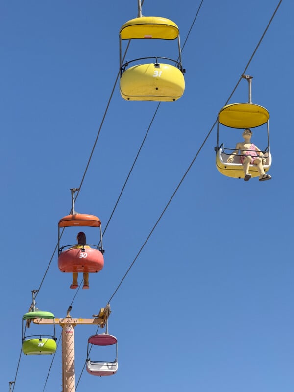 Colorful ski lift chairs, each with a person, suspended high in the air against a bright blue sky. The chairs and cables recede into the distance, creating a sense of depth and motion. The chairs are yellow, red, green, and purple. The scene exudes a playful vibe.