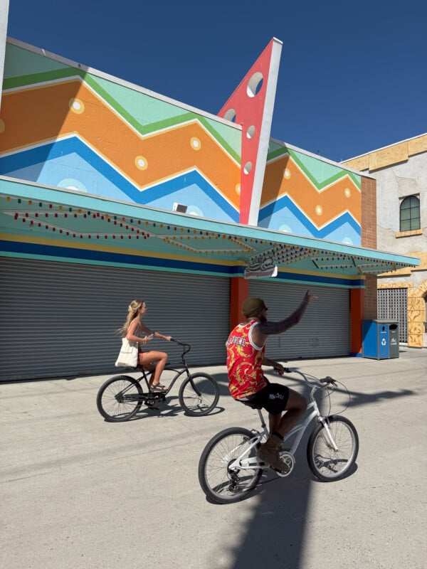 Two people riding bicycles on a sunny day past a brightly decorated building with colorful geometric shapes and a large zigzag pattern. The person at the forefront waves while the other cyclist rides ahead, holding a tote bag.